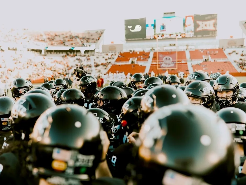 Sports commissioner app team huddle of football players in black helmets on the field before a game.