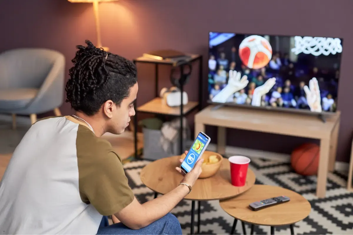 Man using a smartphone app while watching a basketball game on TV in a cozy living room