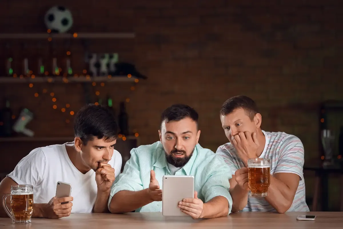 Three men intensely watching a tablet screen with beer mugs, in a sports bar setting
