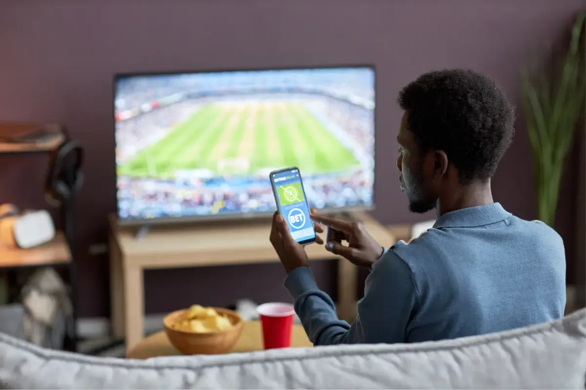 Man using a sports betting app on a smartphone while watching a soccer game on TV