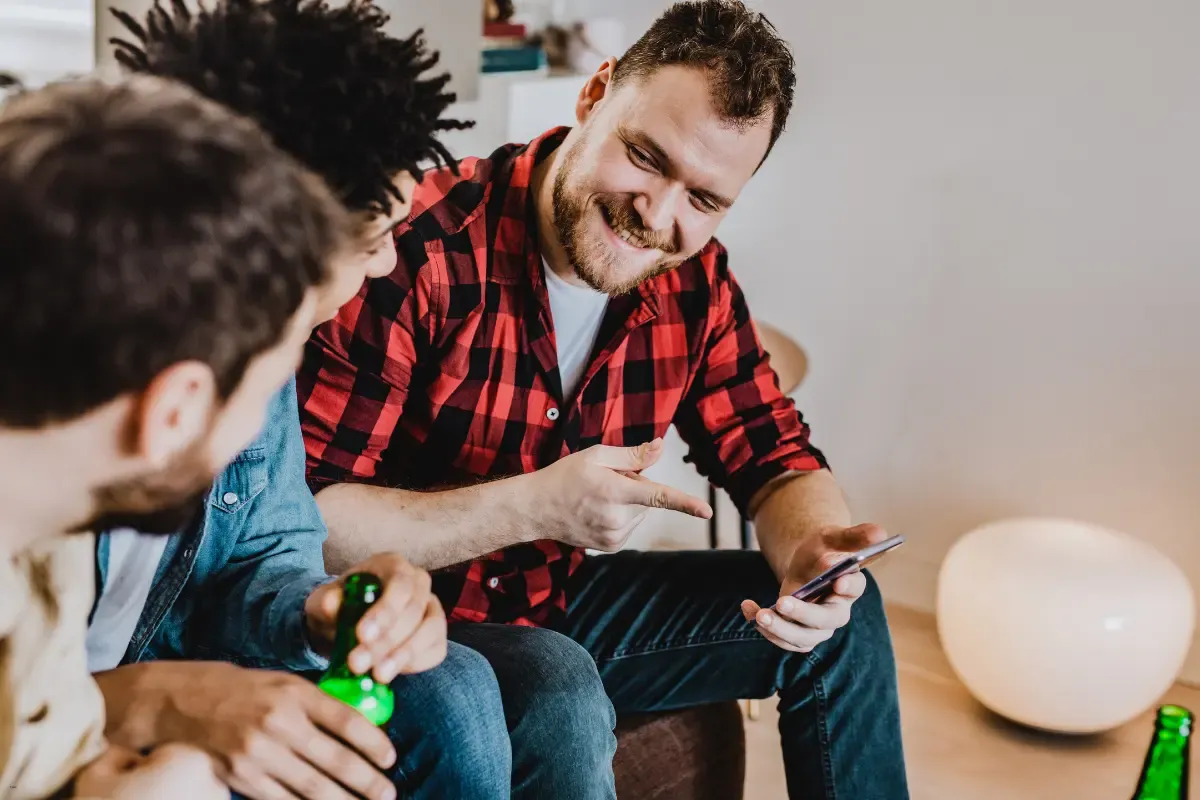 Friends laughing and looking at a smartphone together while holding drinks in a relaxed setting