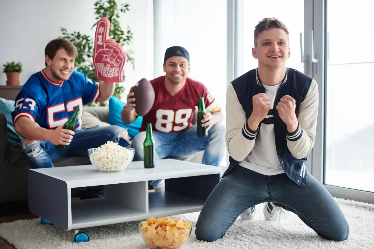 Friends in football jerseys cheering with snacks and drinks, watching a game in a living room