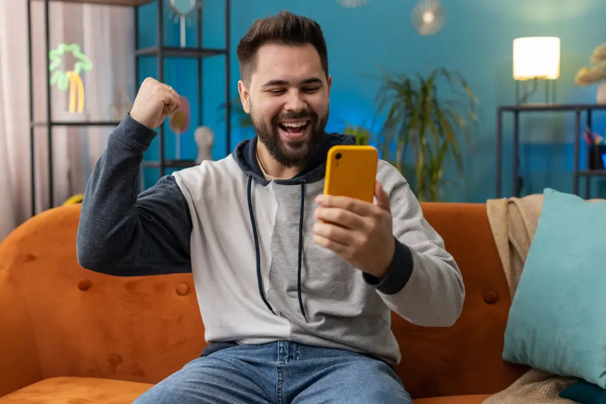 Daily fantasy sports P2P thrill as a man cheers while looking at his smartphone on a cozy orange couch