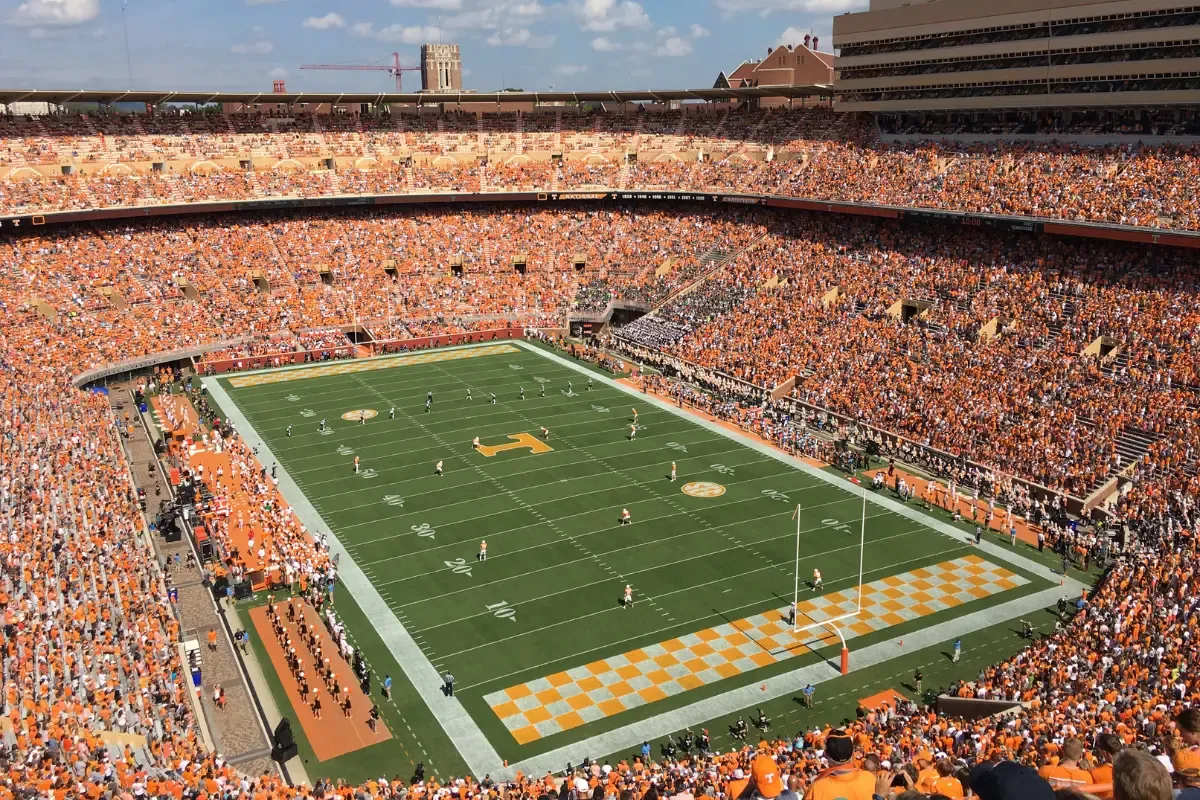Packed college football stadium with fans in orange watching an intense game under a sunny sky