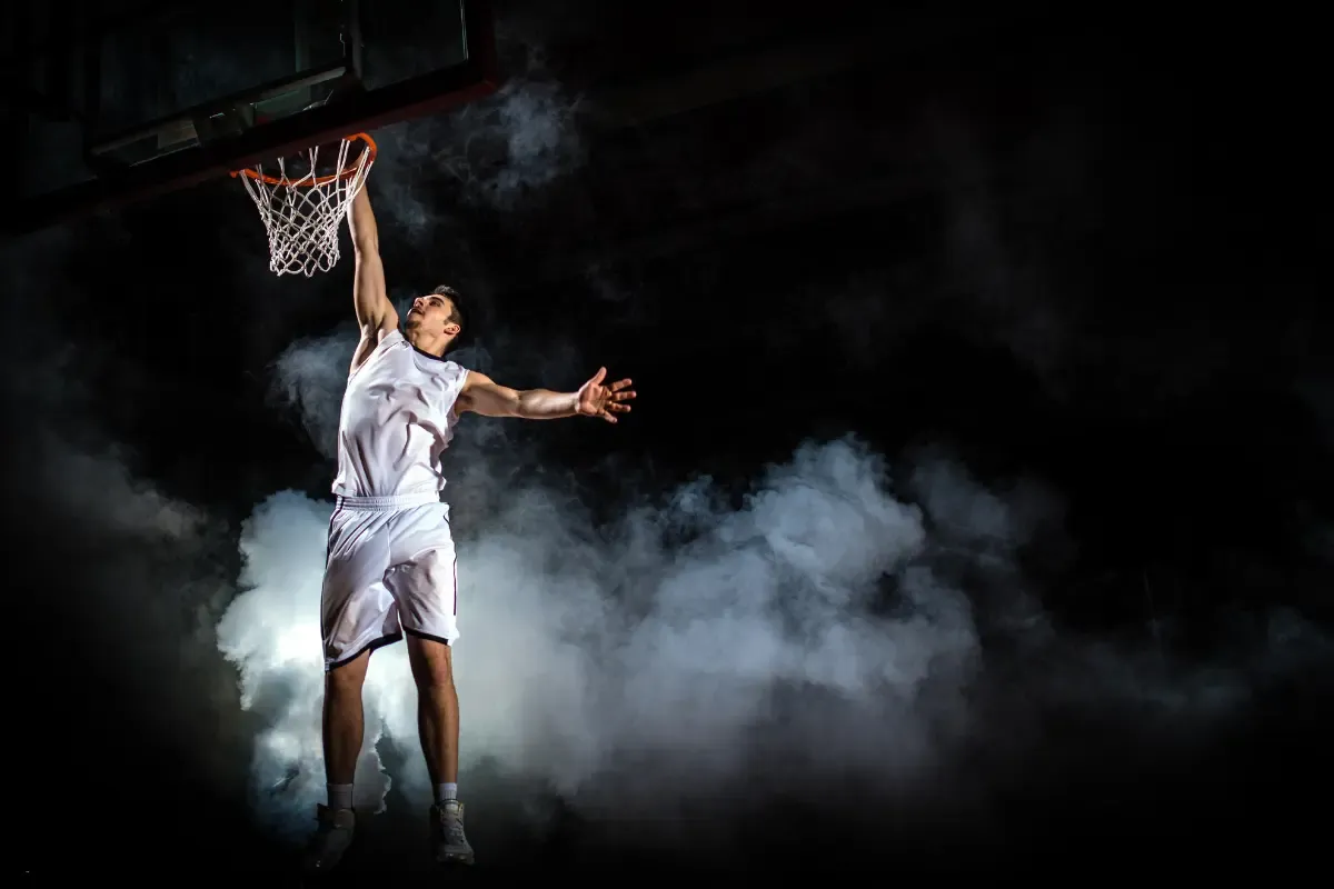 March Madness fantasy basketball player in white jersey leaps for a dramatic slam dunk with smoke in the background