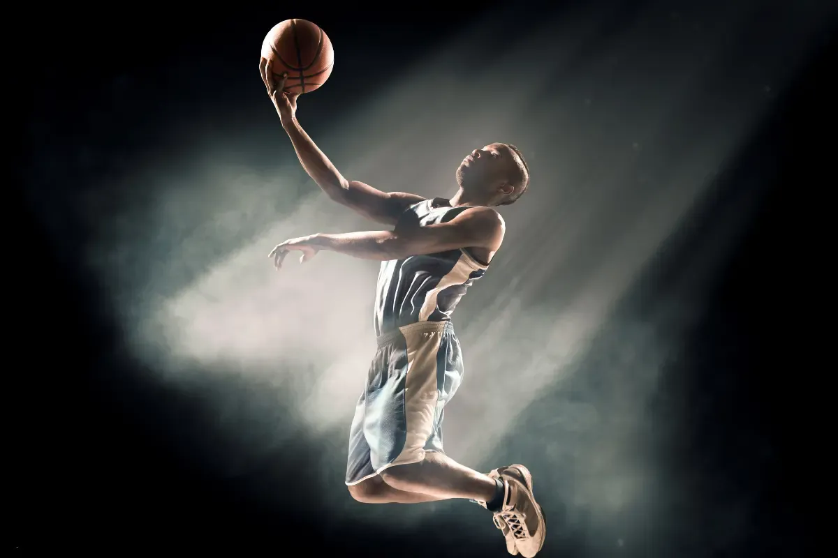 March Madness fantasy player in dark jersey jumps for a layup, illuminated by dramatic spotlight and mist
