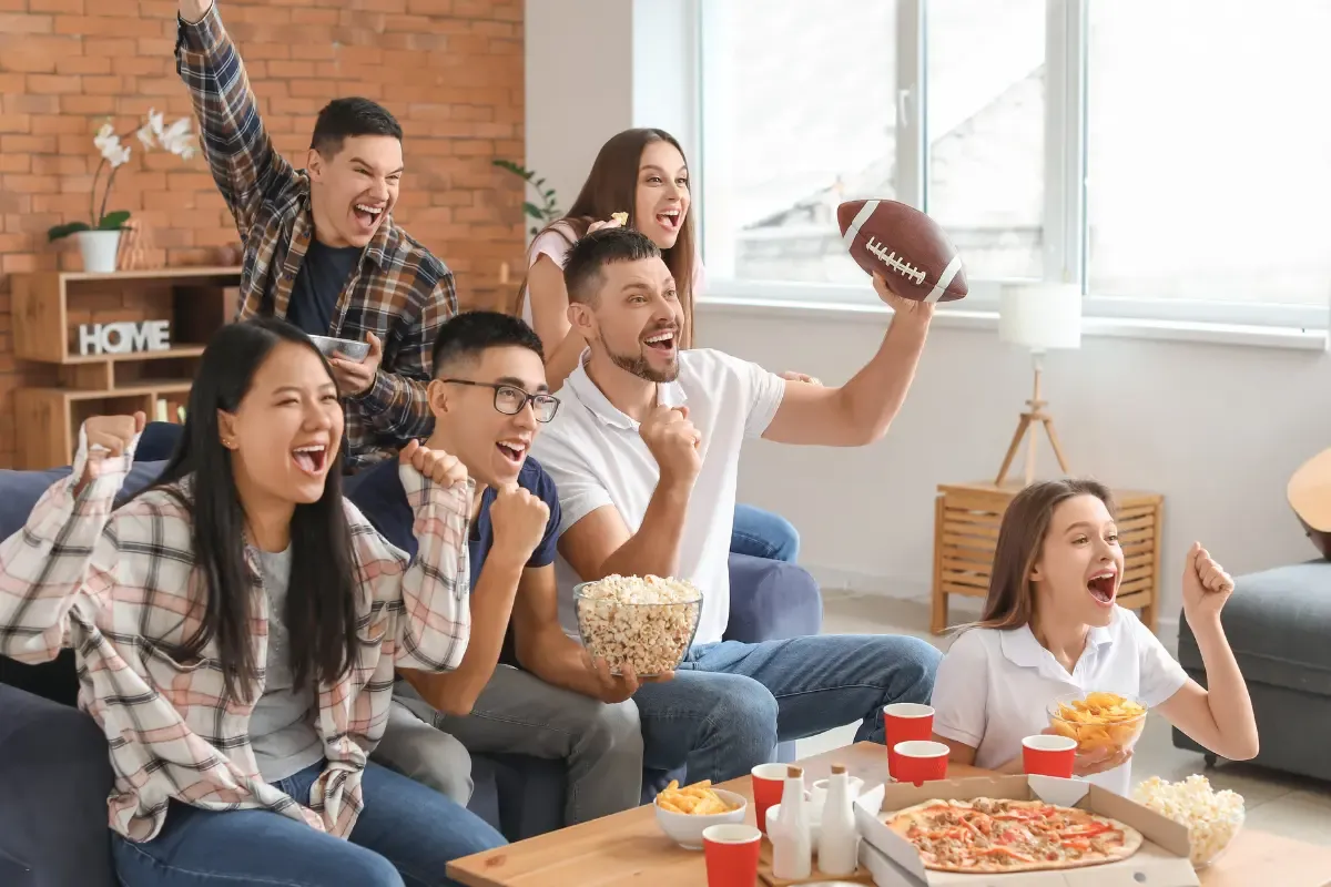 Excited football fans cheering with pizza, popcorn, and snacks during a game day gathering at home