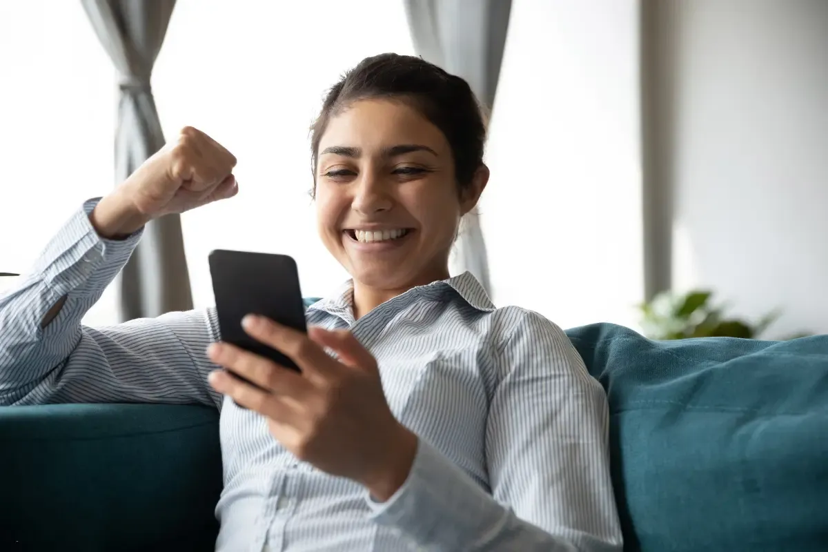Smiling woman raising her fist in excitement while looking at her smartphone on the couch
