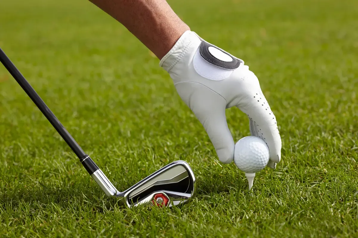 Golfer wearing a white glove placing a golf ball on a tee with an iron club nearby