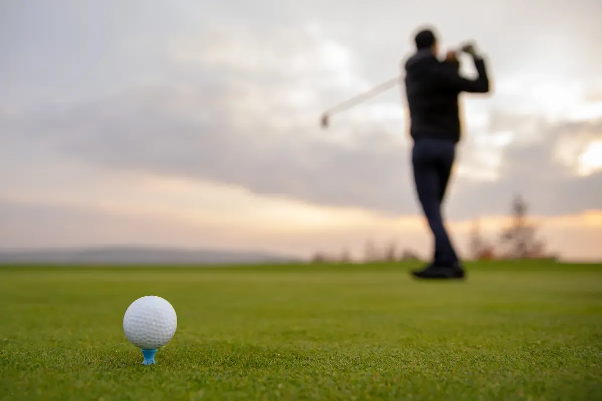 Golf ball on a tee with a golfer swinging in the distance during a cloudy sunset.