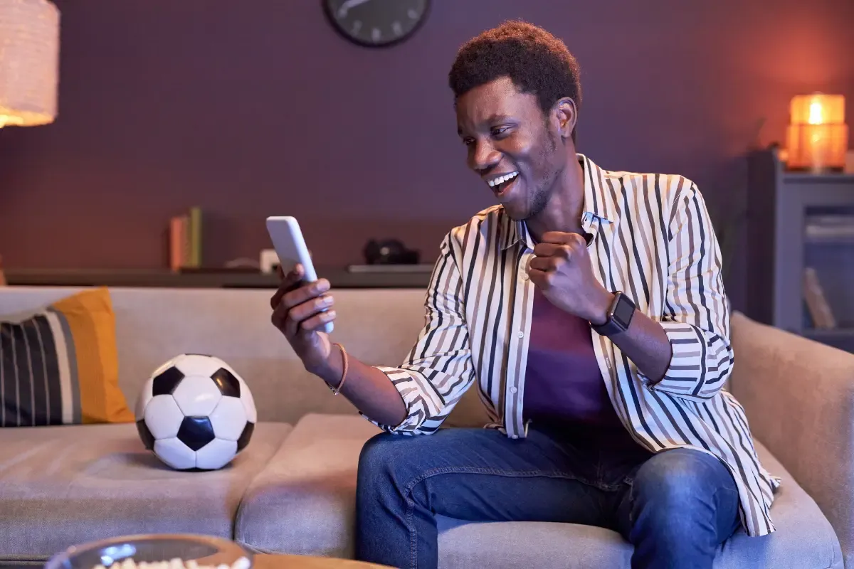 P2P fantasy sports with a man reacting excitedly to his phone while sitting next to a soccer ball on the couch.