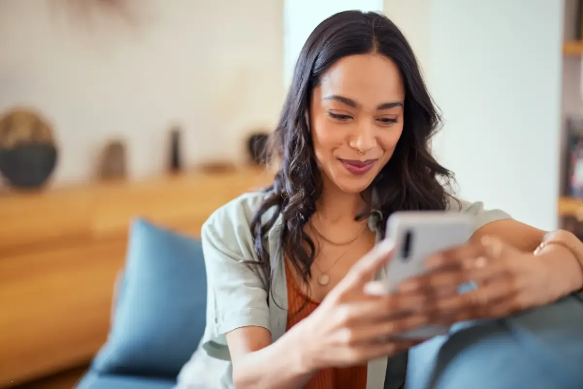 Smiling woman sitting on a couch while looking at her smartphone in a cozy home setting