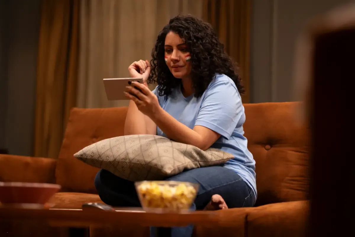 A woman with face paint watching a sports match on a tablet, enjoying popcorn on a brown couch