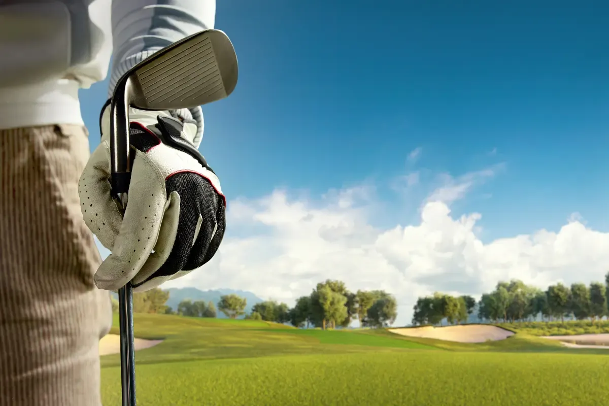 A golfer wearing a glove holding an iron club, overlooking a scenic golf course under a blue sky