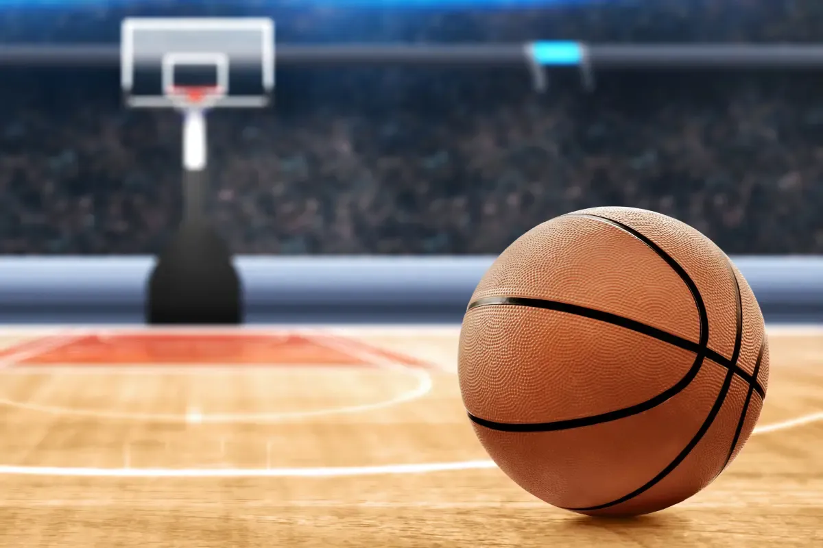 A basketball sitting on the court with a hoop and backboard in the distance, ready for gameplay.