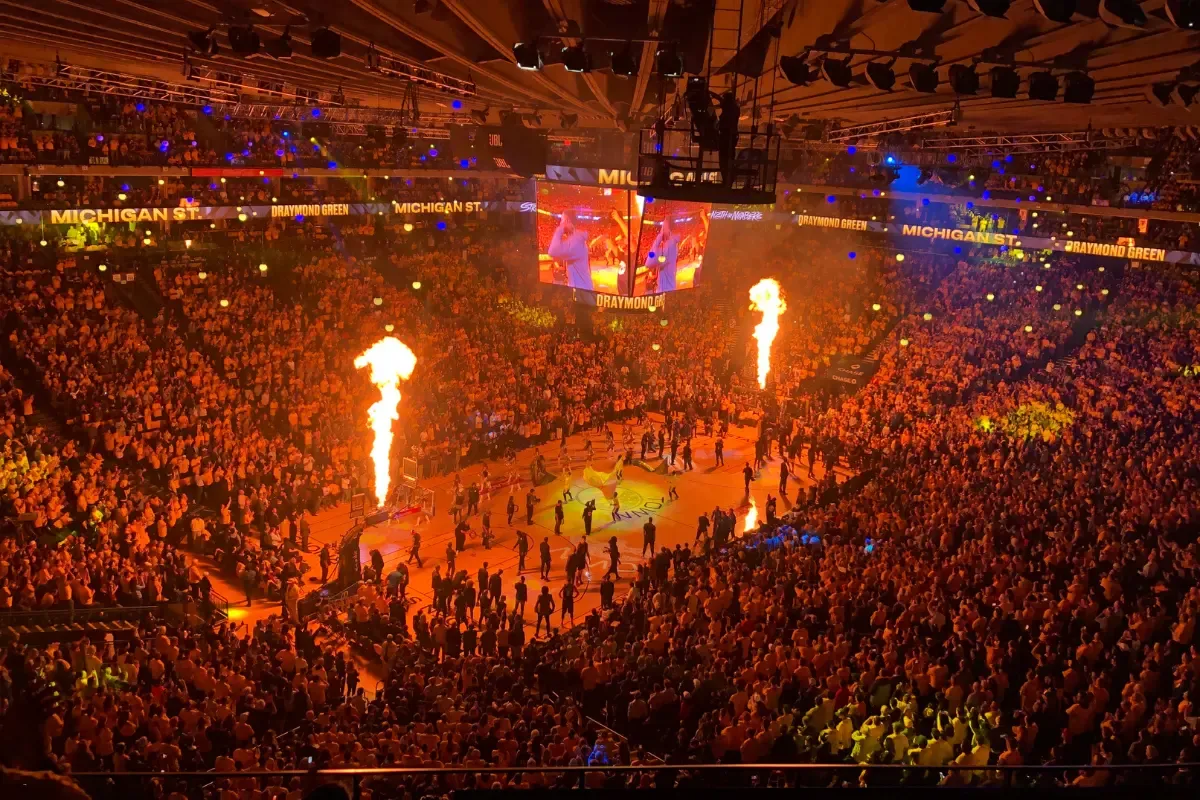 NCAA bracket predictions with a packed basketball arena featuring fire displays and a large screen showing pre-game excitement.