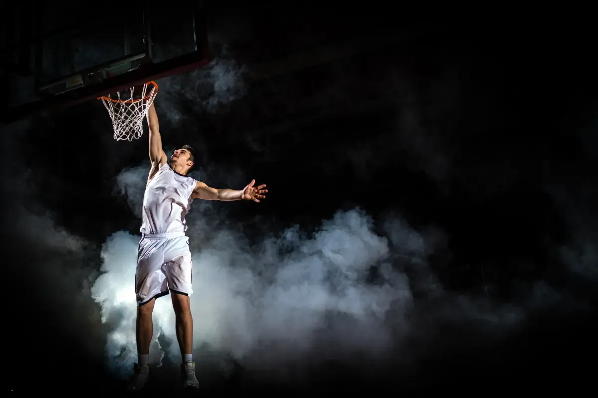 Basketball player in white uniform performing a dramatic dunk through smoke and darkness