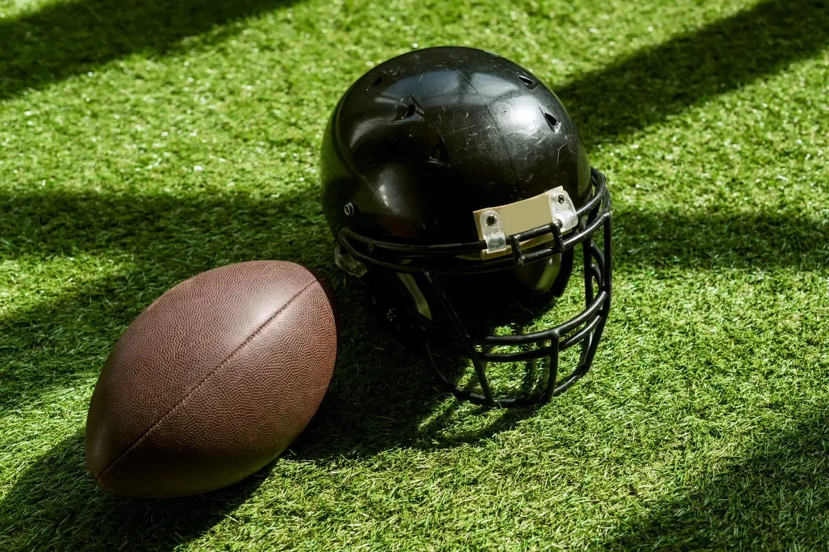 Online NFL pick’em games symbolized by a football and black helmet on artificial turf under sunlight.