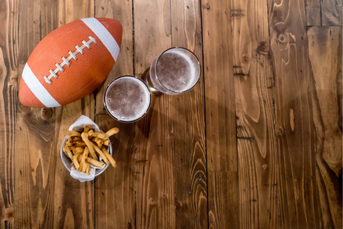 NFL pool strategy symbolized by a football, fries, and two beers set on a wooden bar table.
