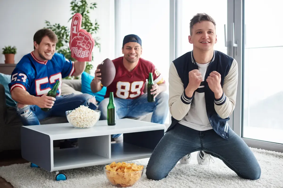 Survivor fantasy league excitement as three football fans celebrate with popcorn, drinks, and foam finger in a bright room.