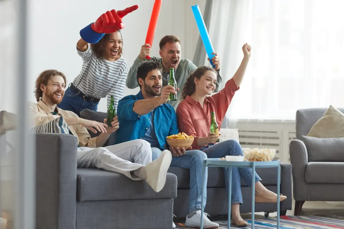 Fantasy league confidence shown by friends cheering with drinks and foam fingers while watching a game at home.