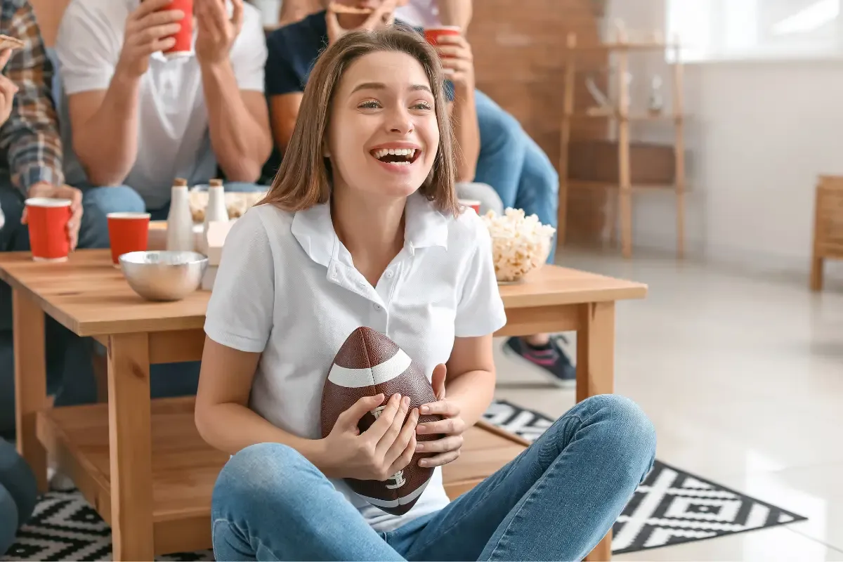 NFL fantasy football excitement as a woman smiles holding a football with snacks and friends in the background