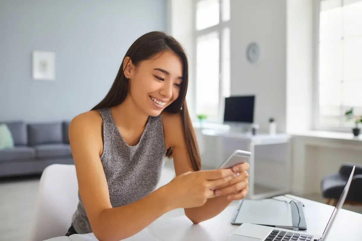 NFL betting fan smiling at phone screen while sitting at a desk with a laptop at home