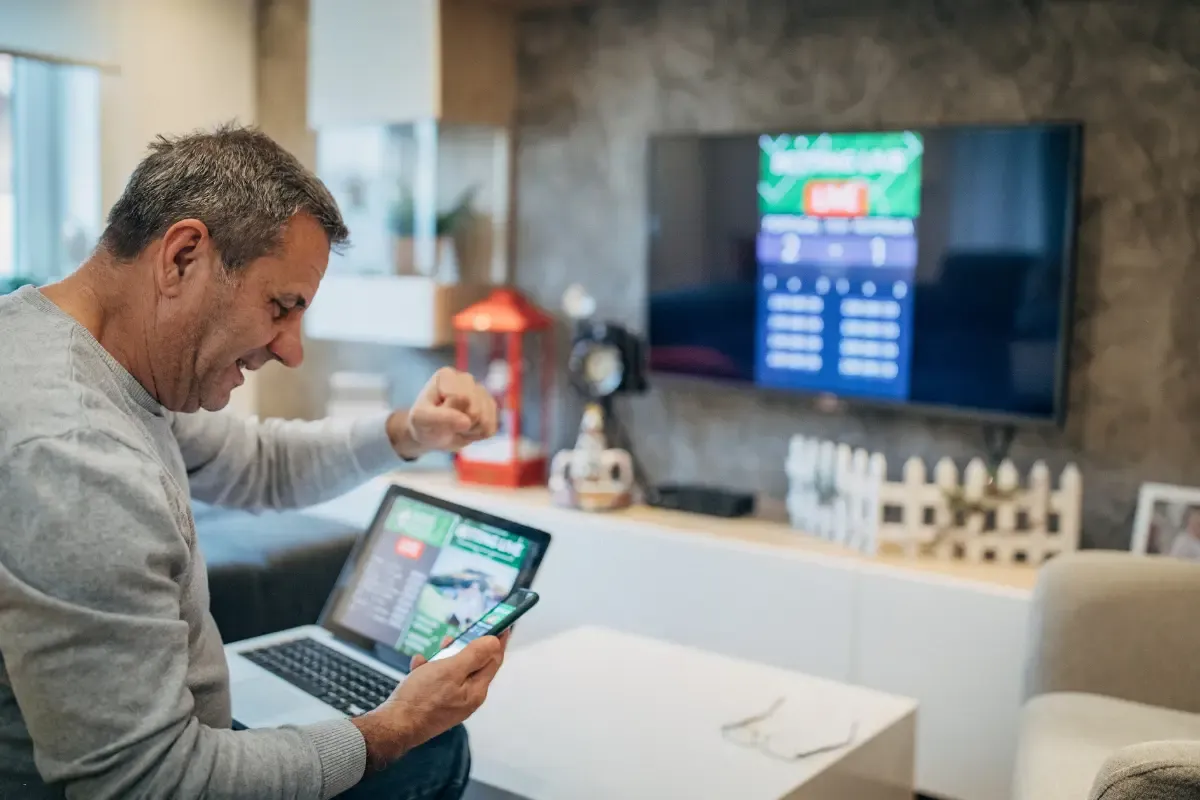 Football pool formats excitement as man watches game results on phone and laptop simultaneously.