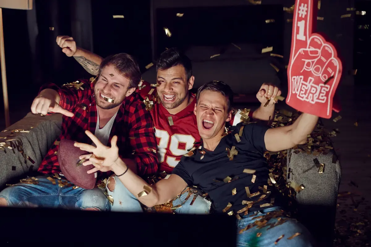 DFS football strategy players watching a game on TV, celebrating wildly with confetti and foam fingers.