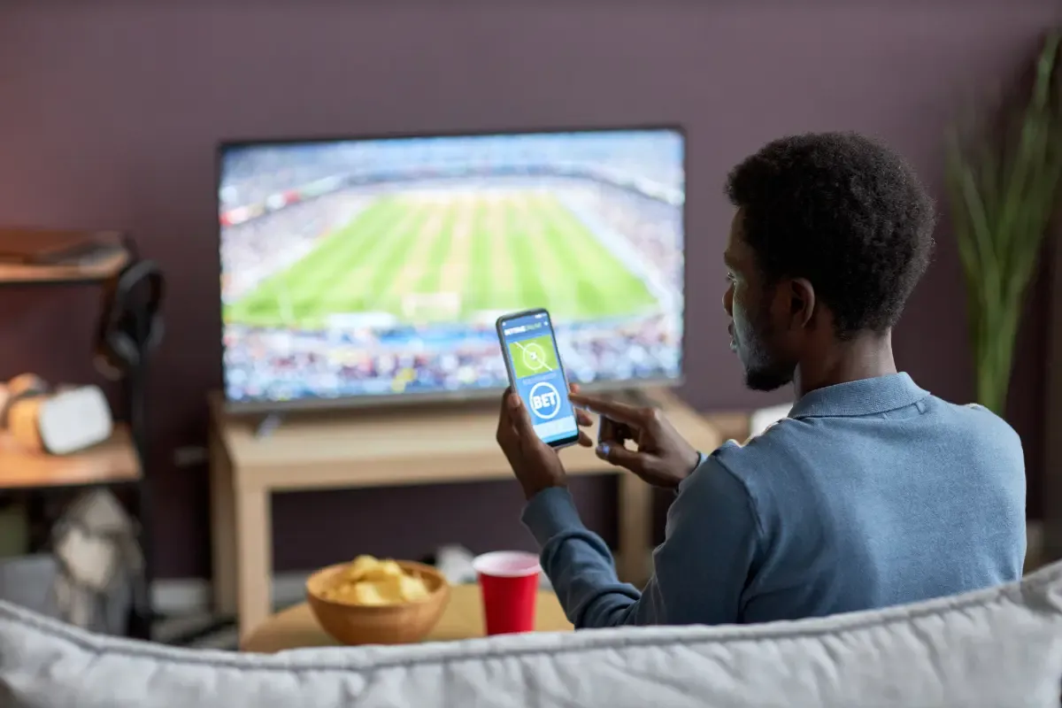 NFL survivor participant placing a bet using a mobile app while watching a live football game on TV.