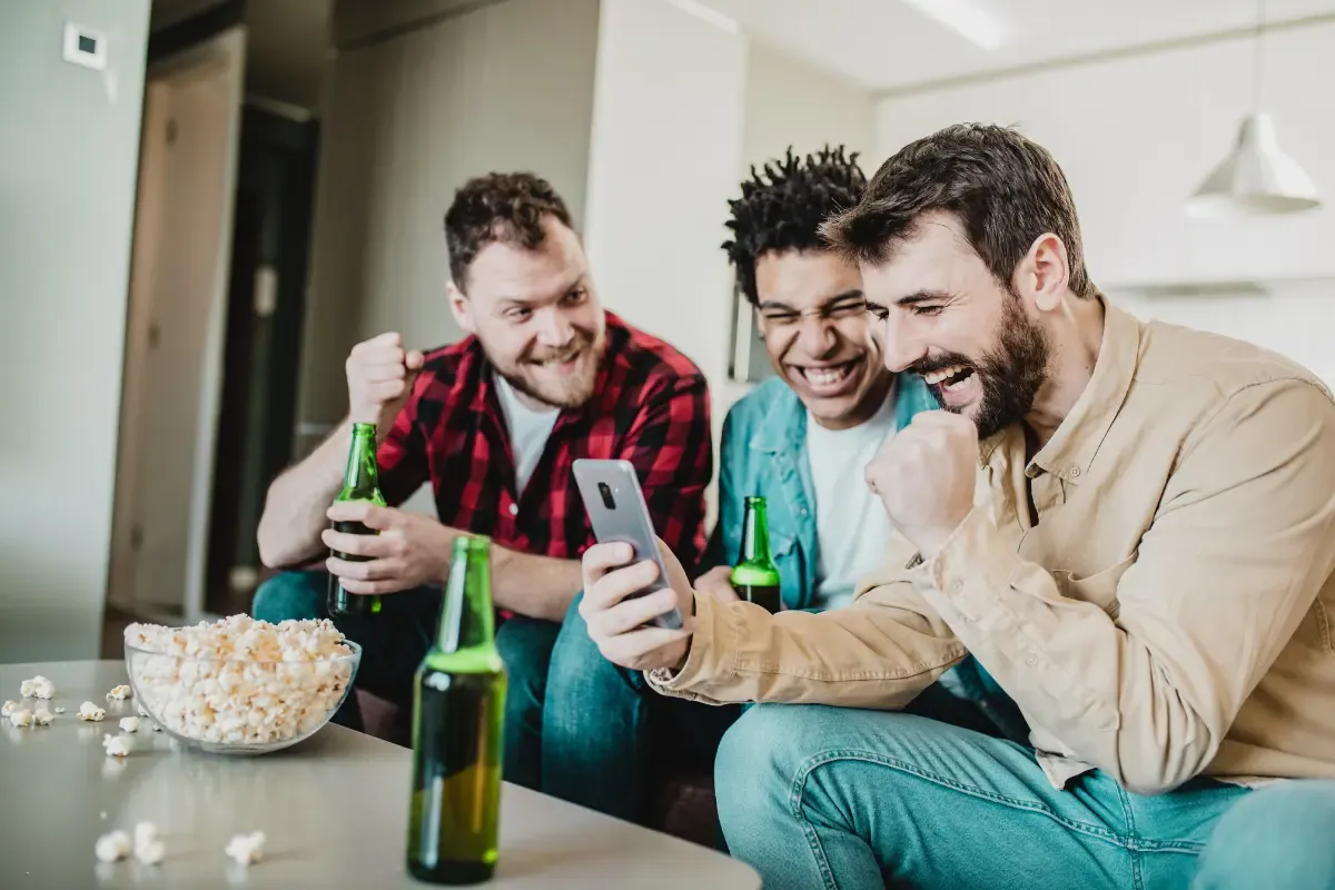 Survivor league football fans cheer while checking game results on a smartphone with drinks and snacks on the table.