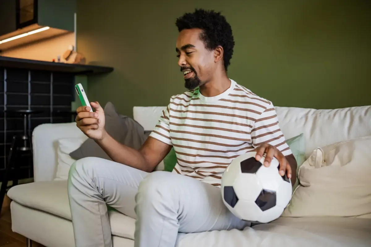 Fantasy betting apps used by a smiling man holding a soccer ball while checking his phone on a couch.