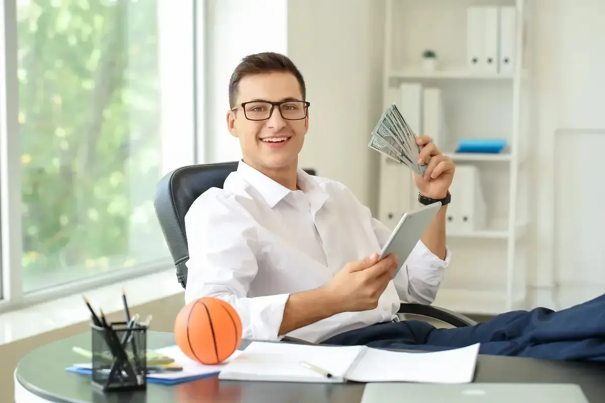 Fantasy league betting success as man smiles holding cash and a tablet in a bright office setting.