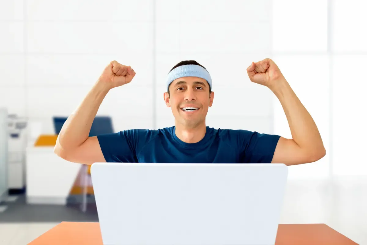 Smiling man celebrating a win while raising his fists in front of a laptop in a bright office setting
