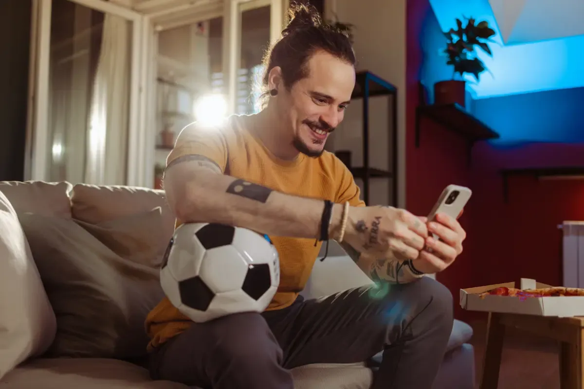 Soccer fan checking his phone while holding a soccer ball on a couch with pizza nearby