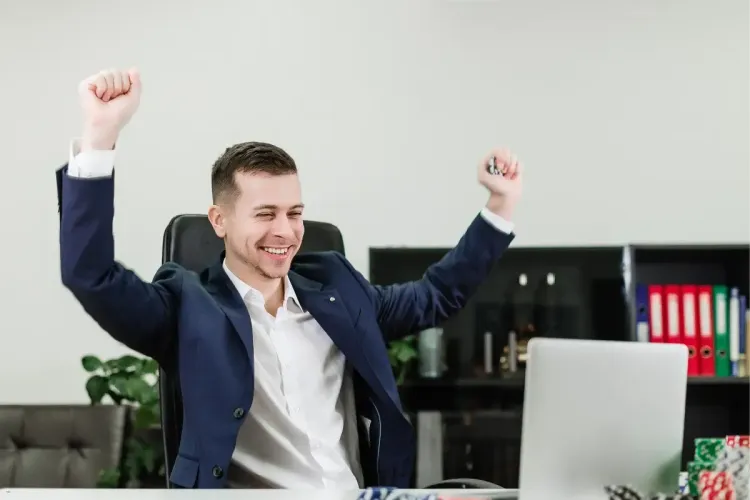 Man in a suit raising his arms in celebration while sitting at a desk with a laptop