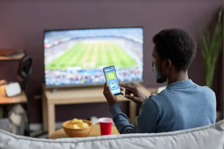Peer-to-peer fantasy sports gameplay as a man checks his smartphone while watching a live soccer match on TV.