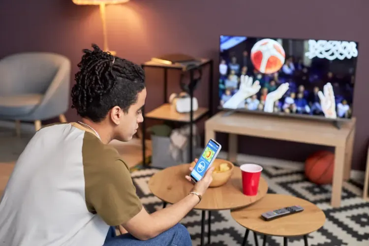 DFS roster building featuring a man analyzing a sports app on his phone while basketball plays on the TV nearby