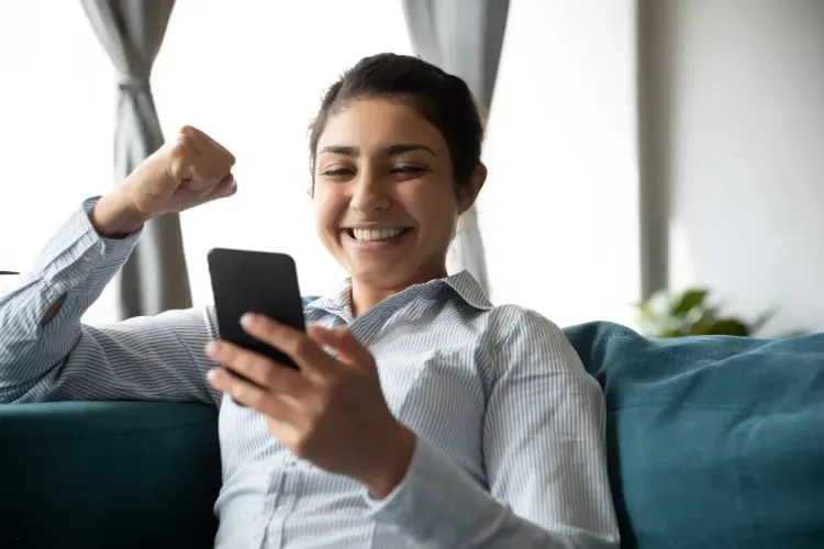 Smiling woman raising her fist in excitement while looking at her smartphone on the couch
