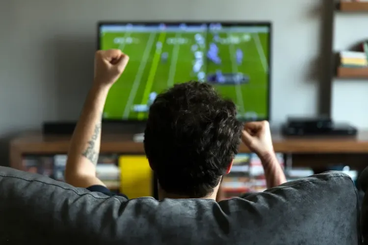 A football fan cheering while watching a game on TV from a cozy living room couch.