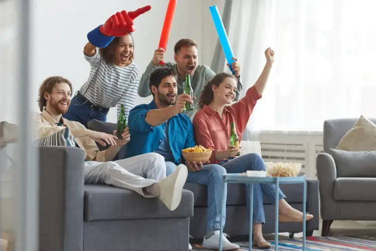 Fantasy league confidence shown by friends cheering with drinks and foam fingers while watching a game at home.